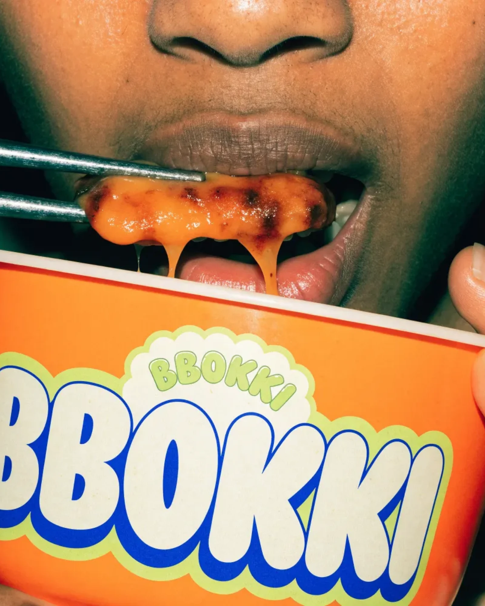 BBOKKI, close-up of a mouth biting glossy rice cakes from a cup