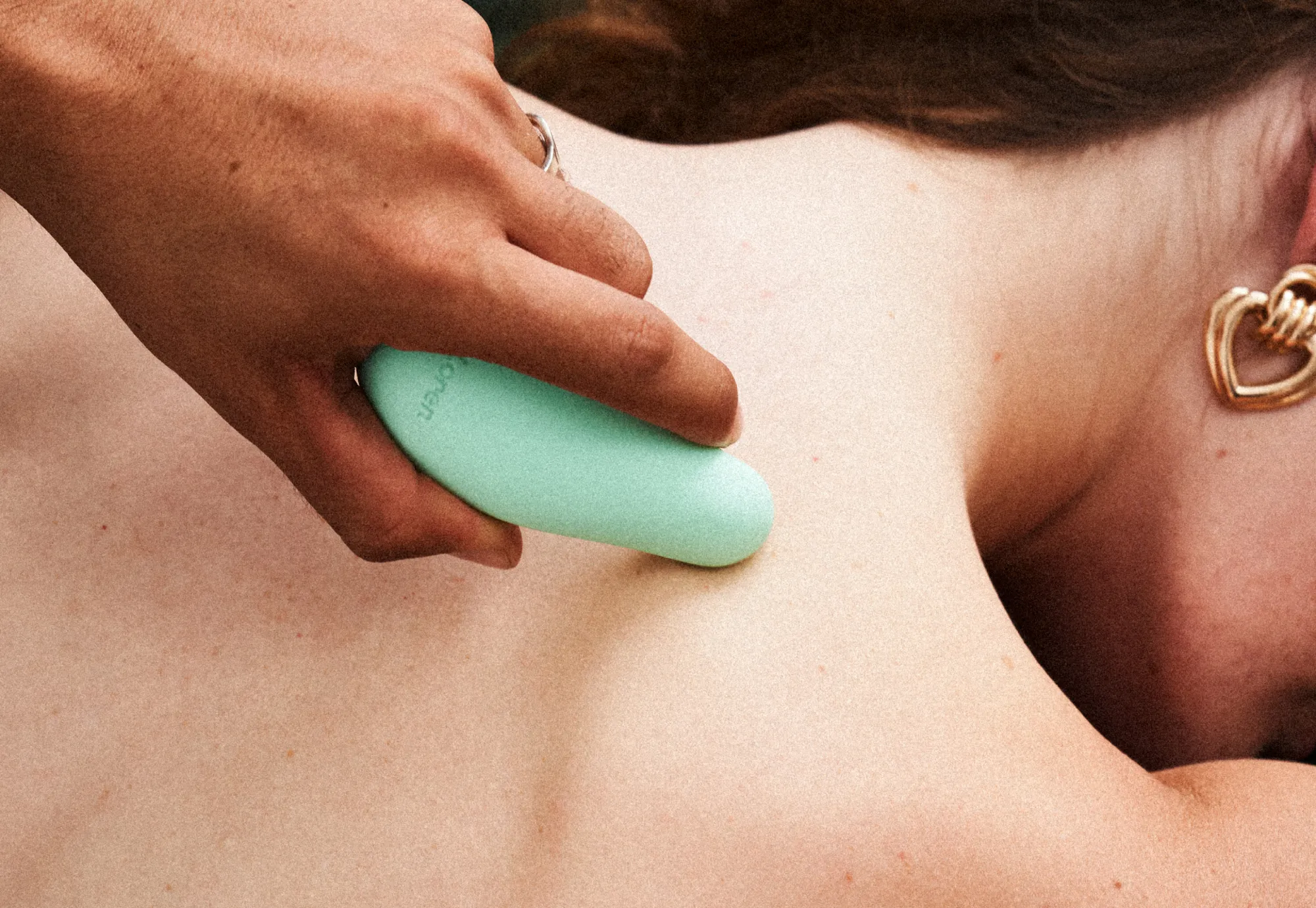 LOREN, a hand pressing a mint massager into a bare shoulder beside a gold heart earring