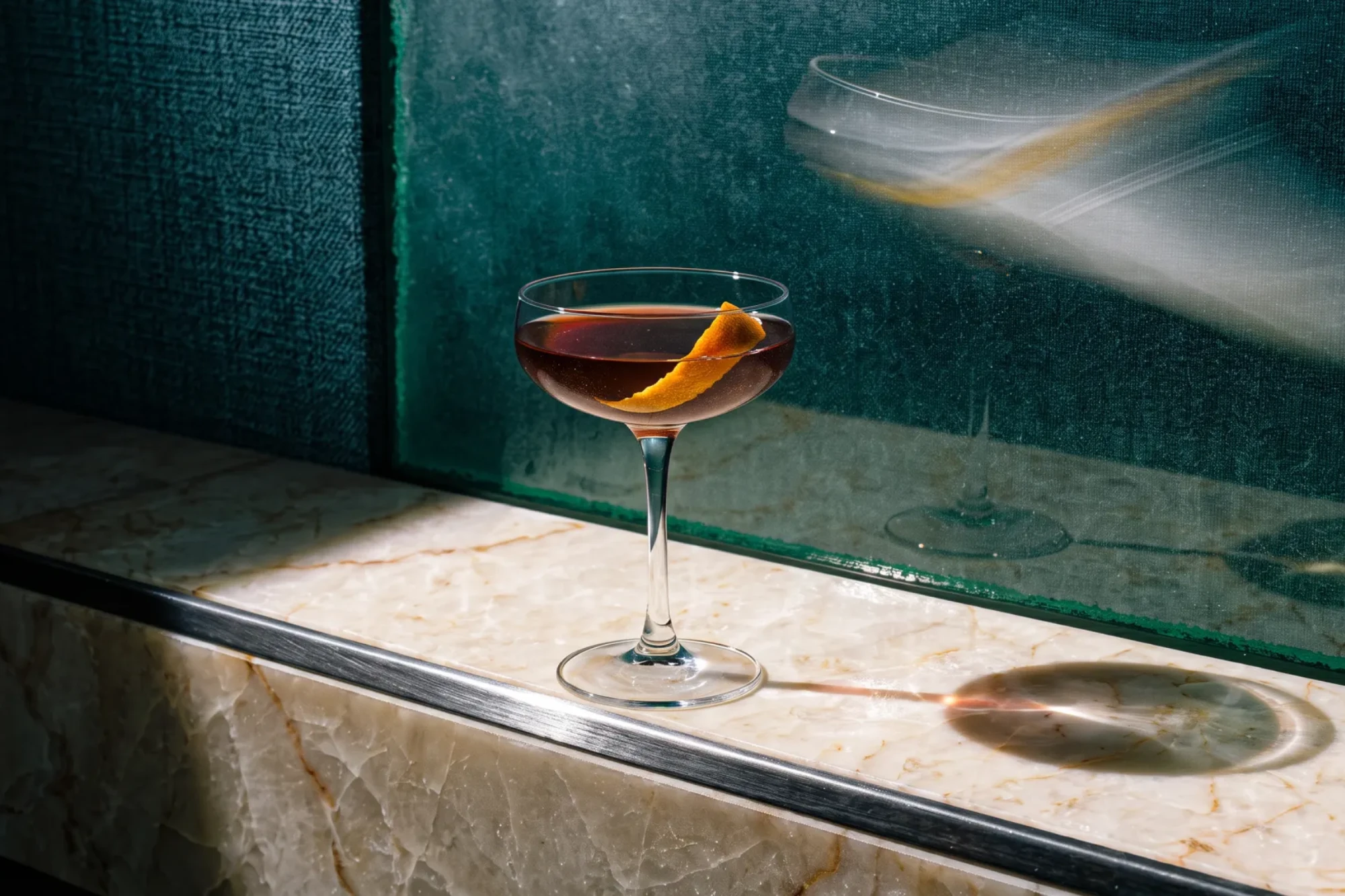 Picada, a cocktail with an orange twist on a green marble ledge in hard afternoon light